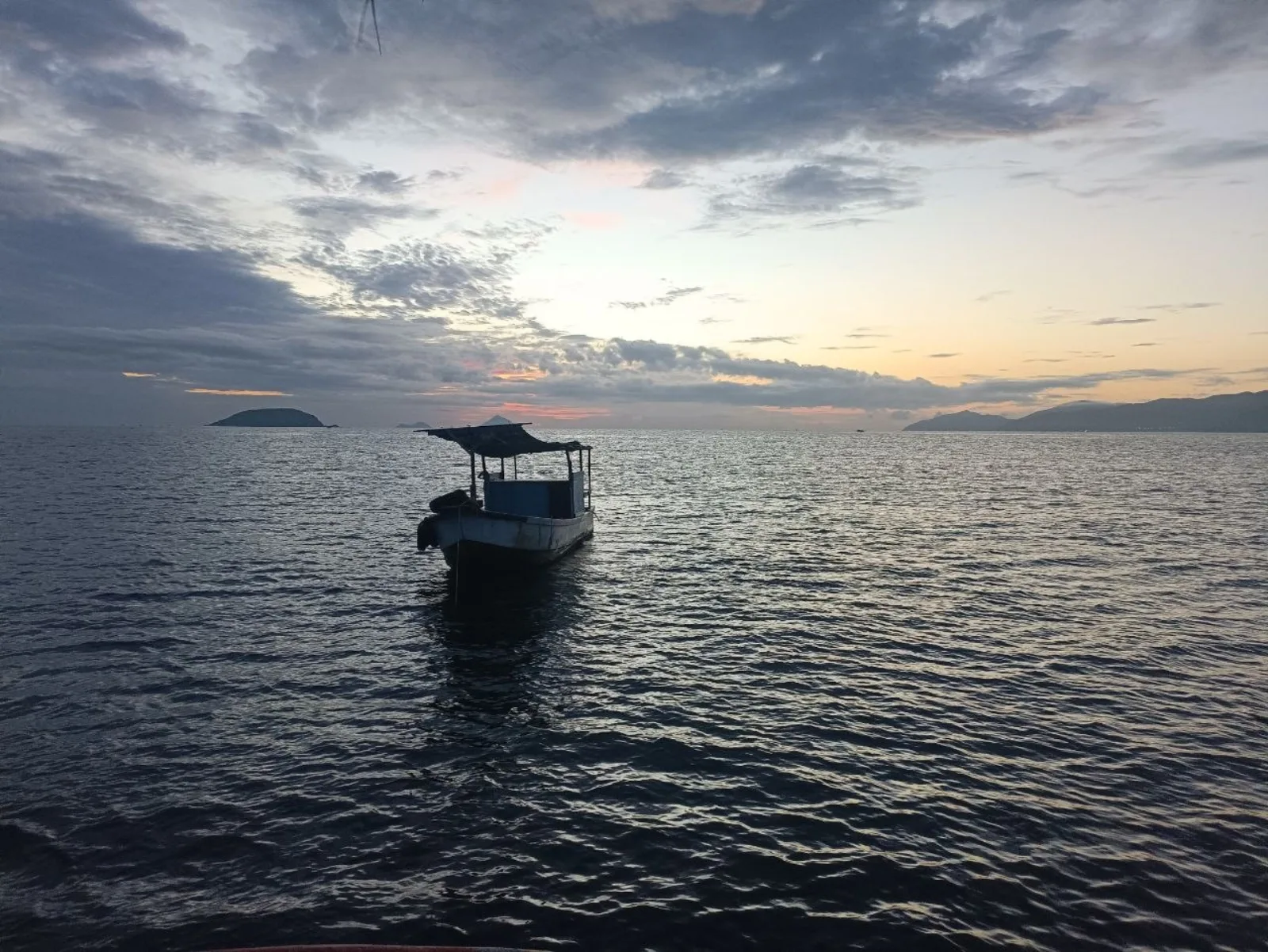 Небольшая лодка с навесом стоит на морской глади в сумерках, на горизонте видны силуэты островов и гор под облачным небом